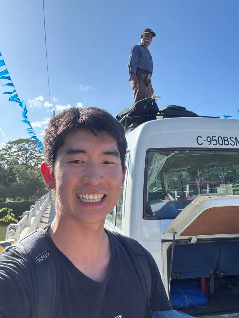 Selfie of me smiling while the bus driver climbs on top of the bus to move bags
